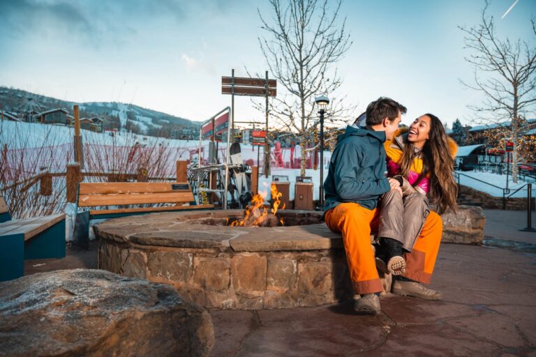 A couple enjoys a cozy moment by a fire pit, with a stunning mountain backdrop at sunset.