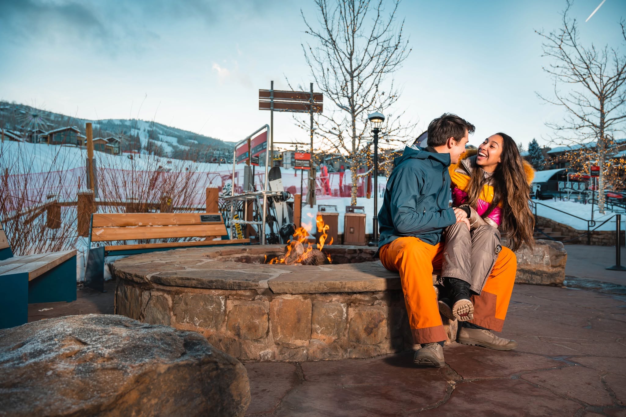 A couple enjoys a cozy moment by a fire pit, with a stunning mountain backdrop at sunset.