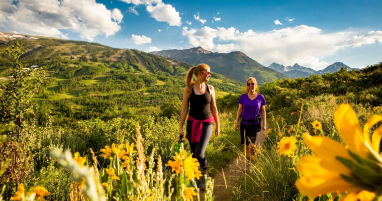 Two women hikers stroll through a sunlit alpine meadow of yellow wildflowers, with green mountain peaks and dramatic clouds in the background.