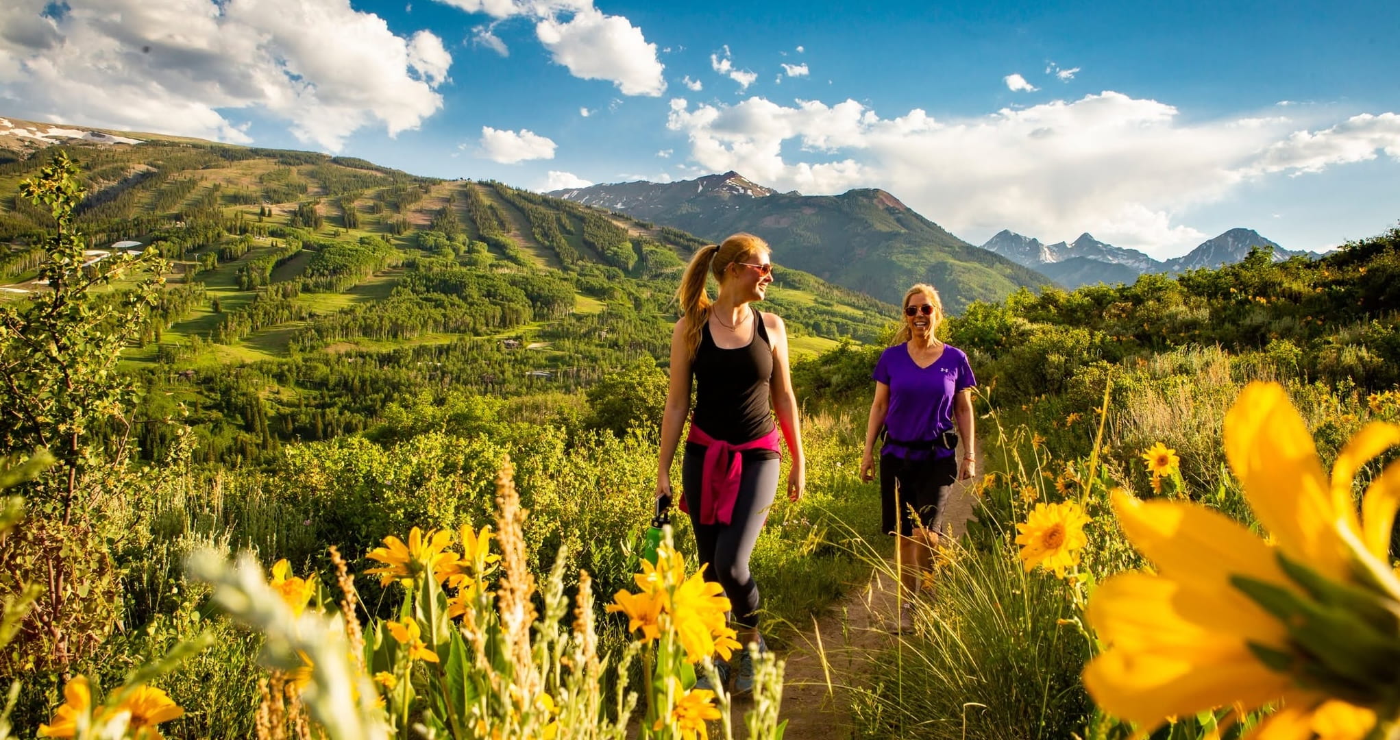 Two women hikers stroll through a sunlit alpine meadow of yellow wildflowers, with green mountain peaks and dramatic clouds in the background.