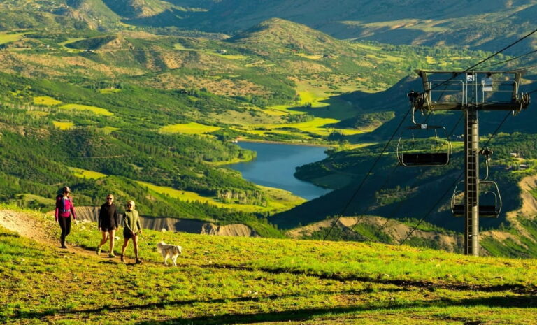 A group of people walks a dog on a mountain trail, with a ski lift visible in the background.